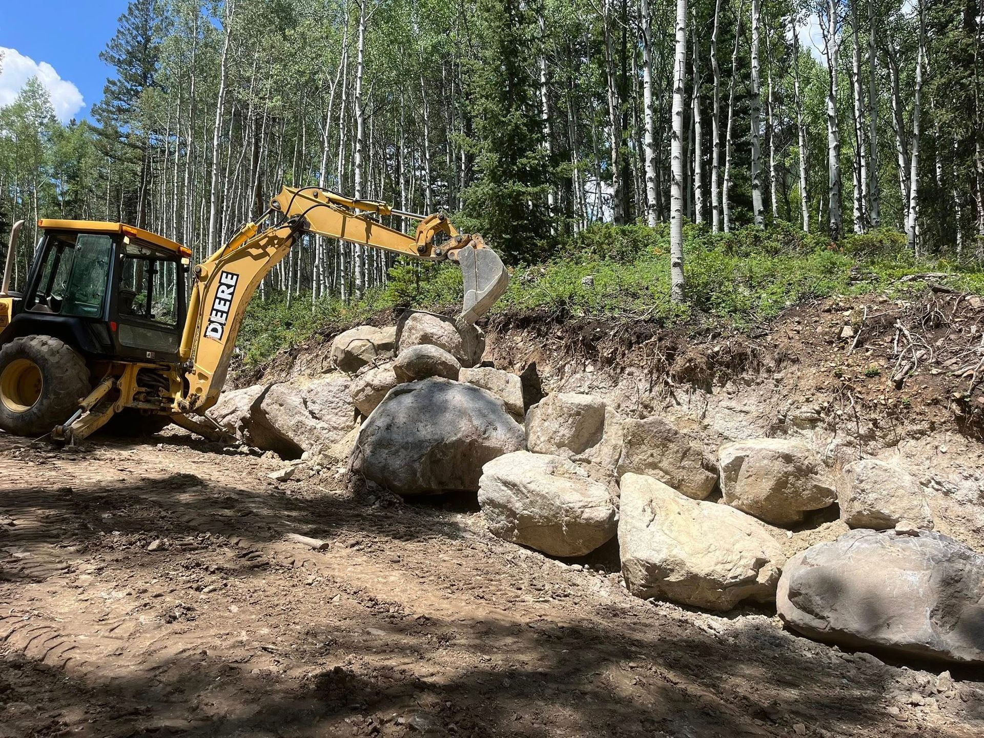 Yellow excavator moving large boulders on a dirt slope in a forest clearing