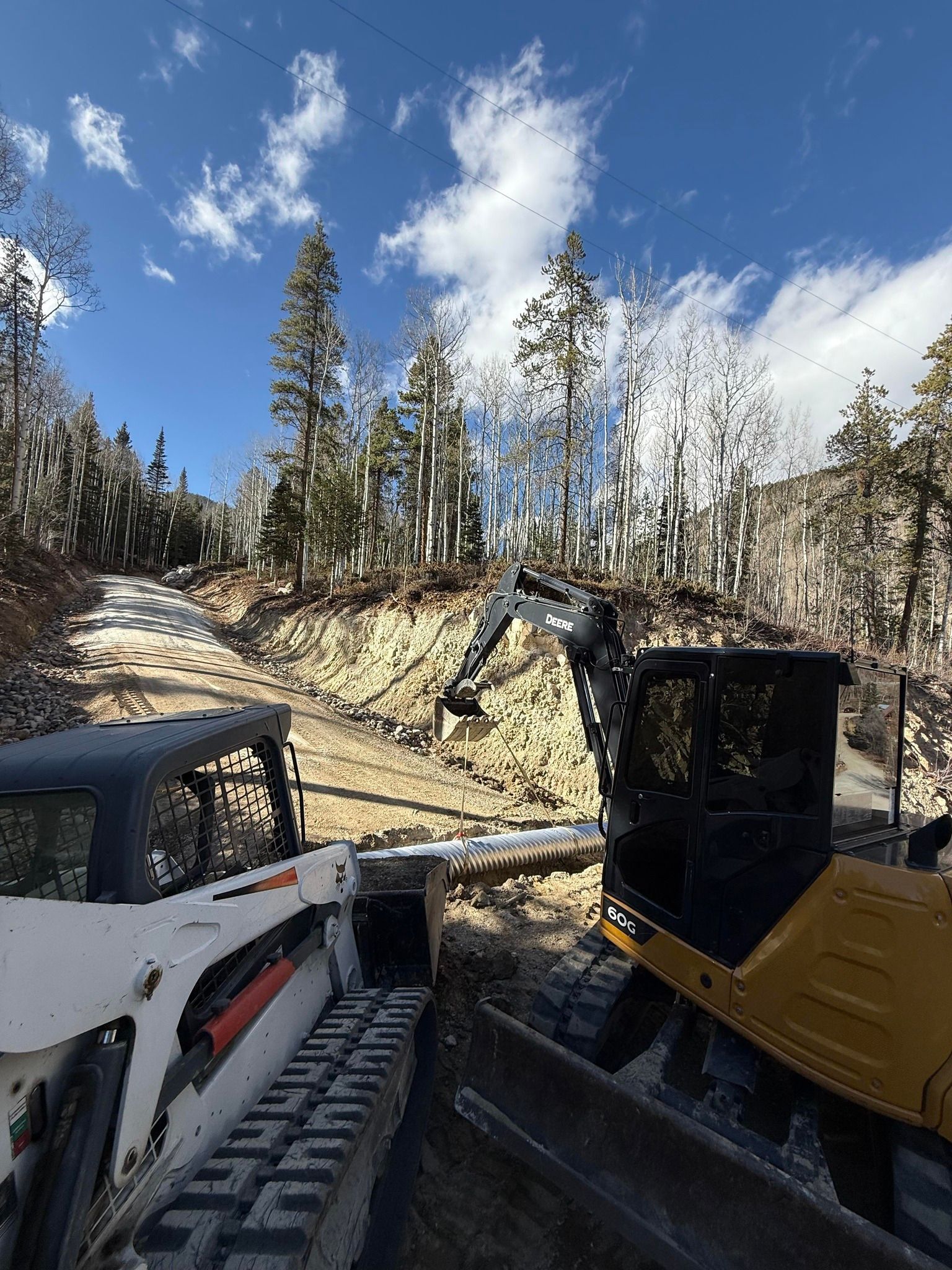 Two construction machines on a rocky forest slope under a blue sky with clouds.