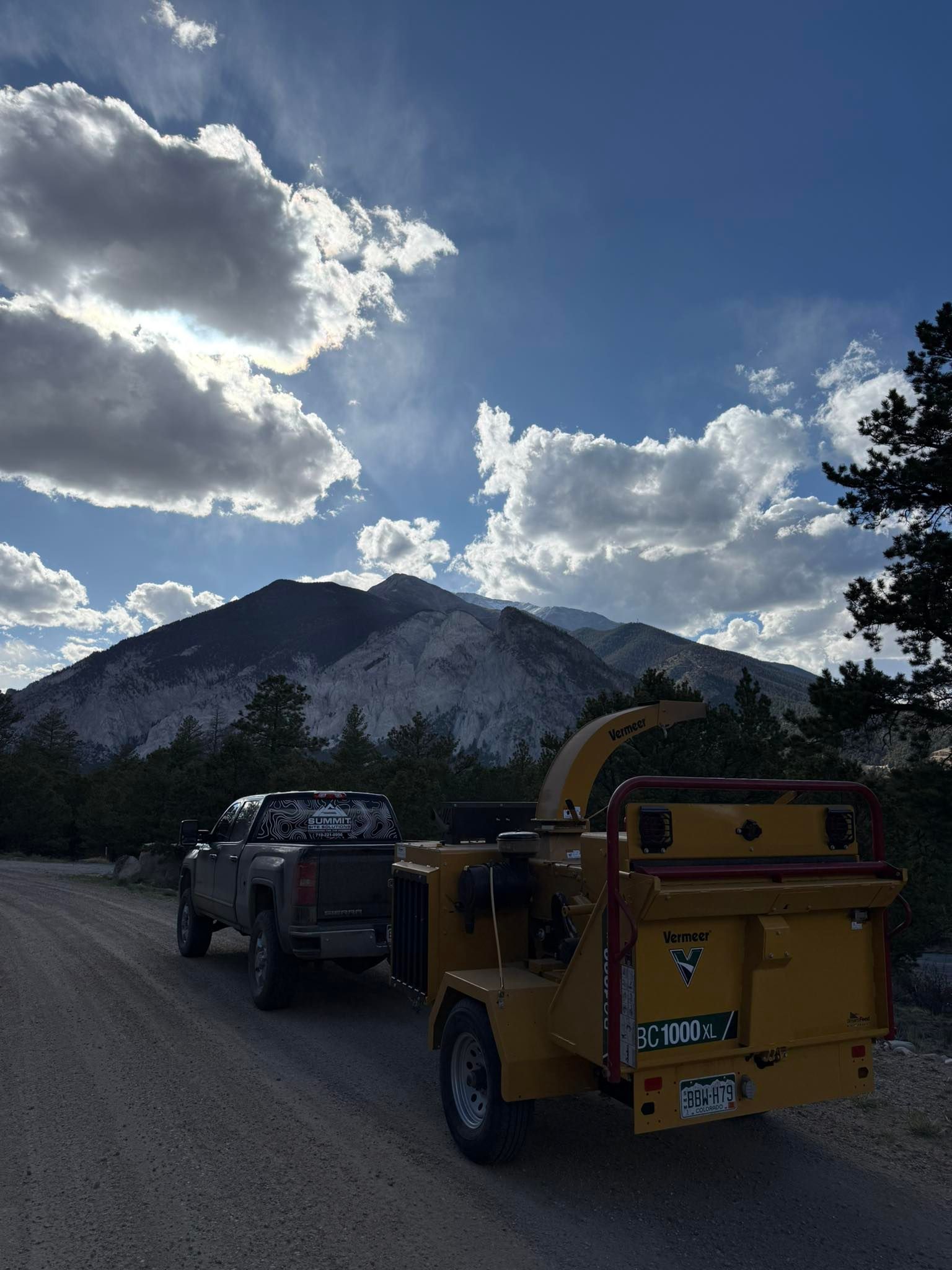 Yellow wood chipper truck towing a trailer on a gravel road beneath cloudy mountain skies