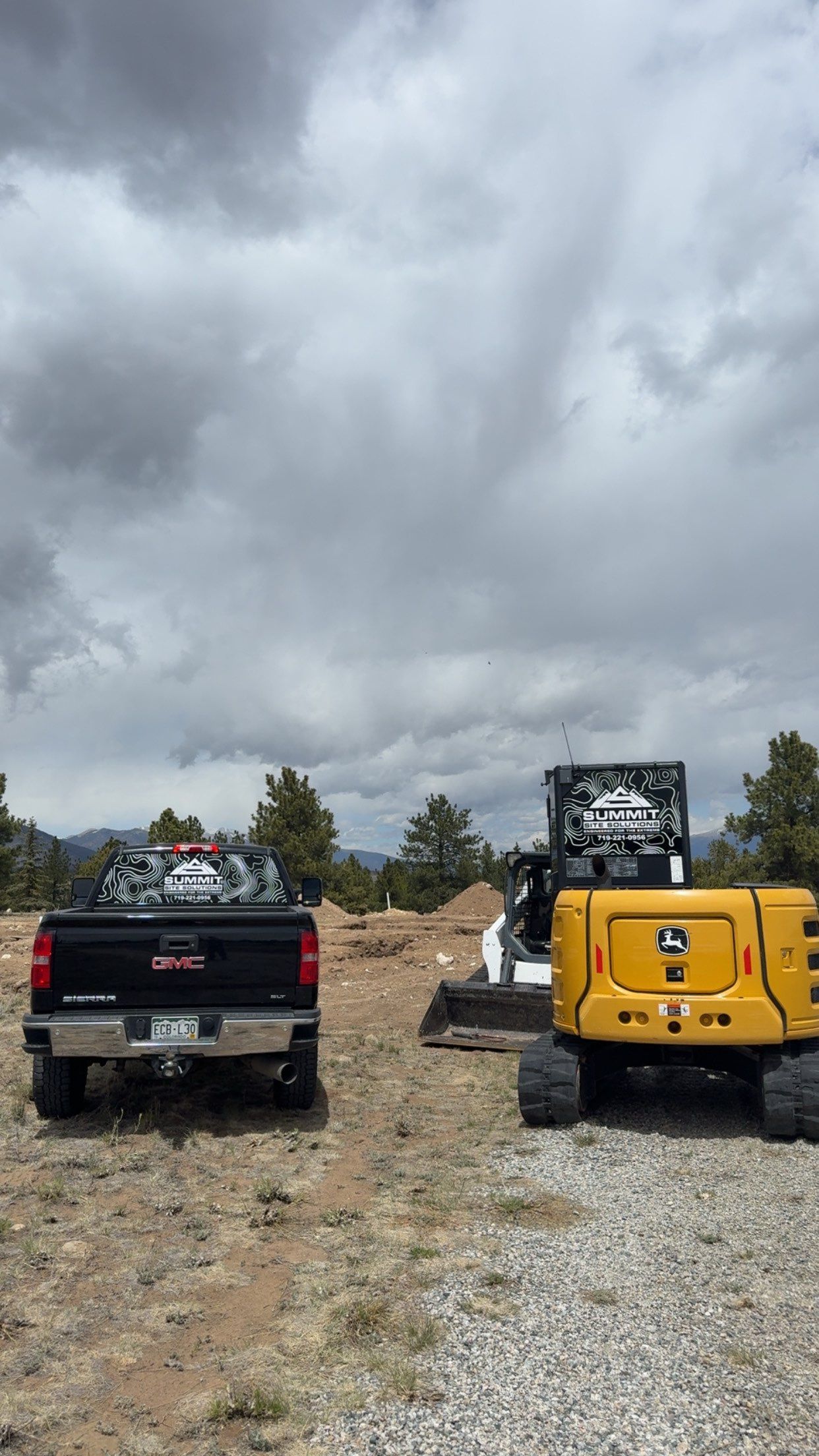 Two off-road trucks parked on a gravel trail under a cloudy sky, with trees and brush in the background.