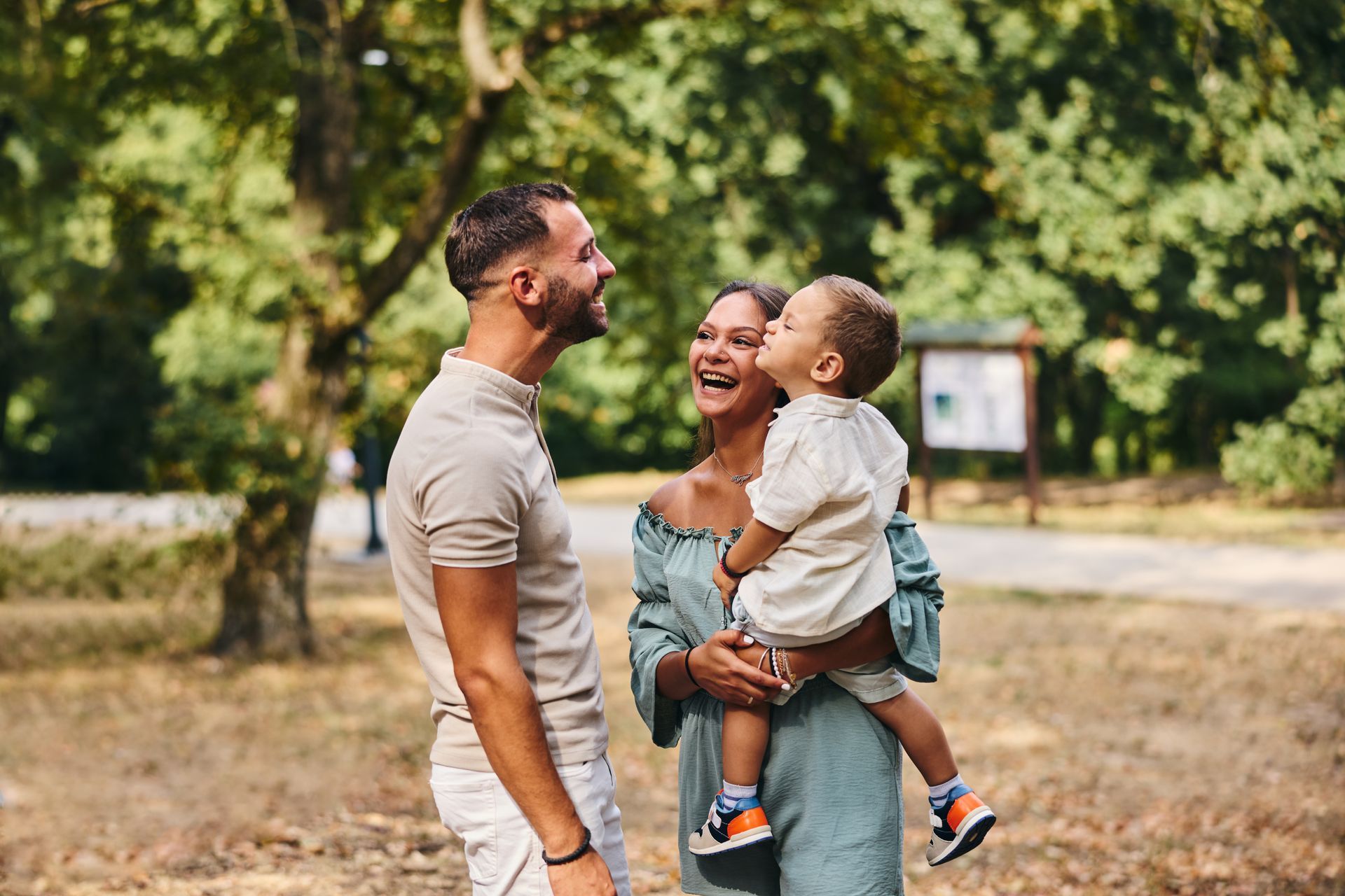 Family laughing in a park; mother holding child who kisses her cheek, father smiling.
