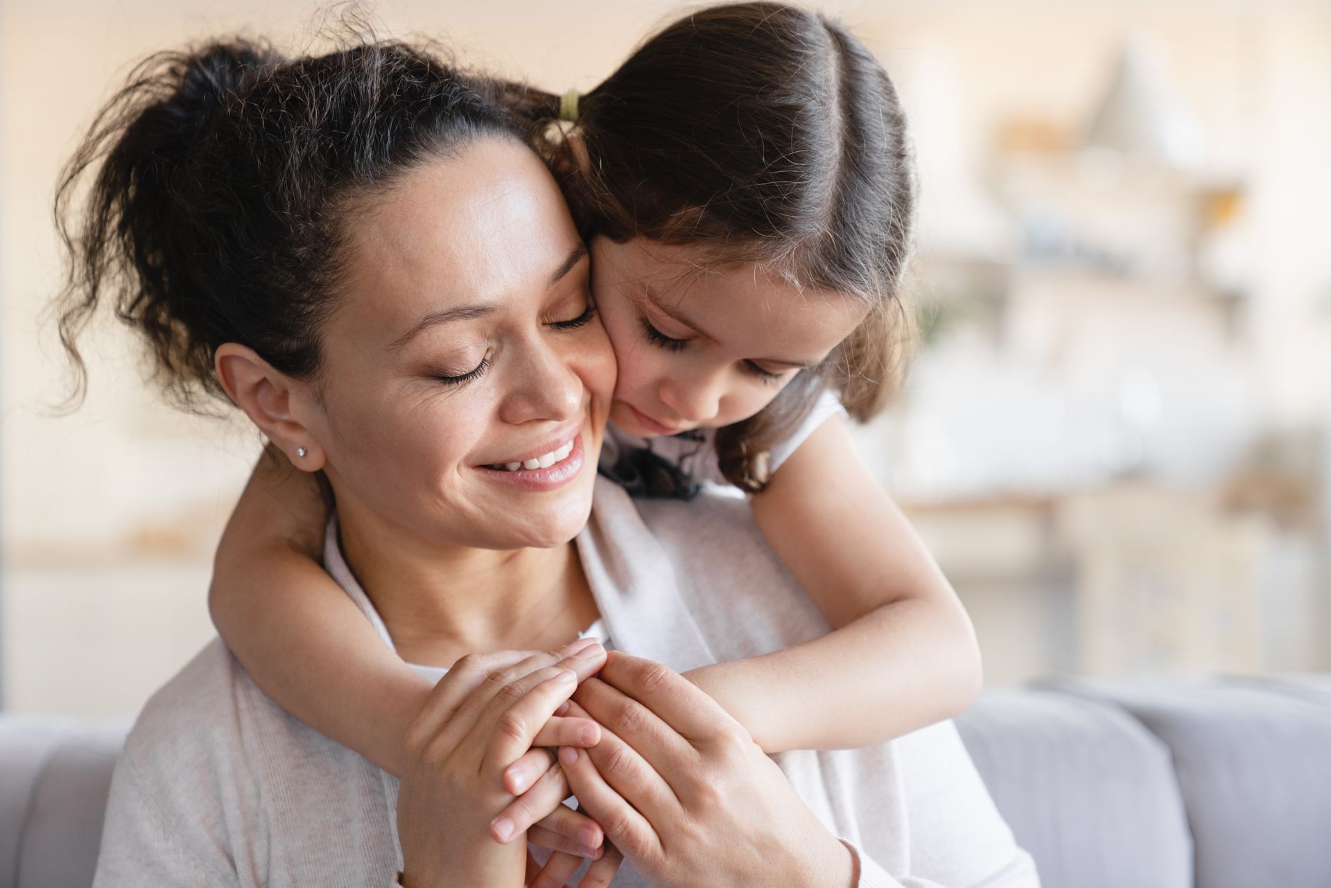 A child hugs a woman from behind, both smiling, indoors.