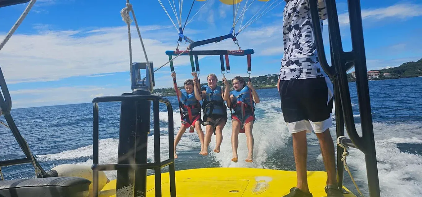 Three people parasailing over the ocean, towed by a boat on a bright day. The parasail is white and yellow against a blue sky.