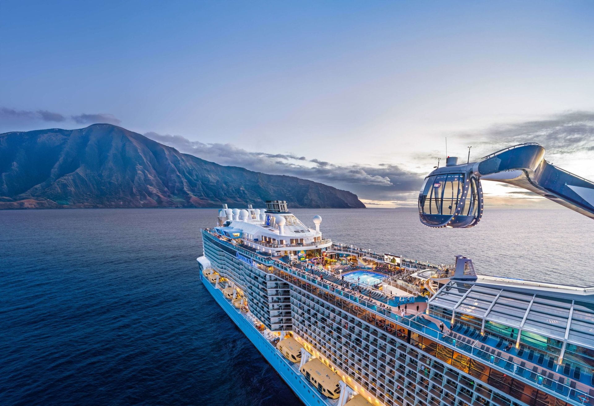 A large cruise ship at sea with a gondola-like structure extending towards the deck, mountains in the background.