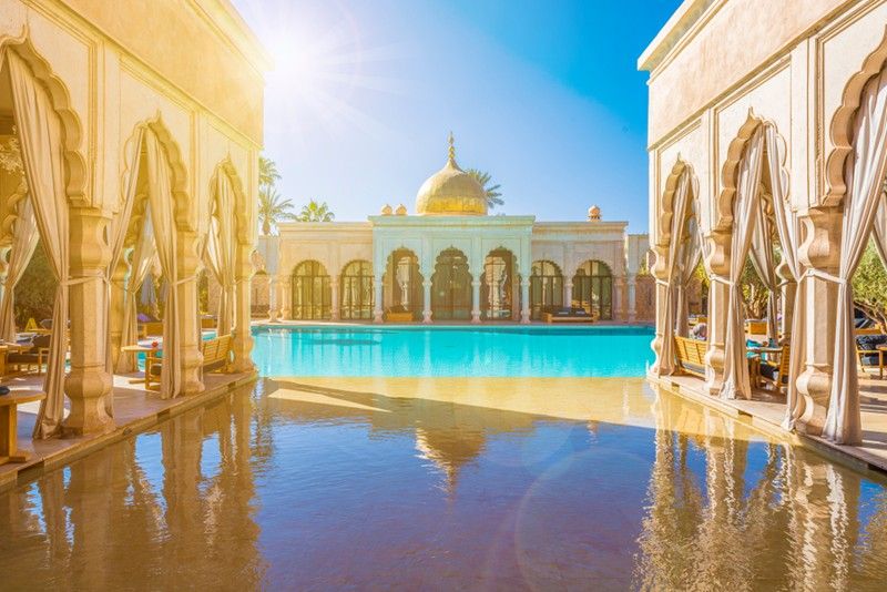 A luxurious Moroccan-style resort with a golden-domed building reflecting in a large turquoise pool, under a bright sunny sky.