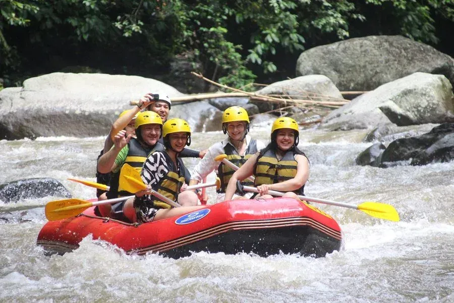 People in a red raft, wearing yellow helmets and life vests, white water rafting on a river with rocks and trees.
