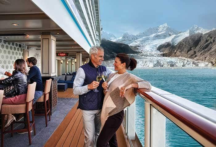 Couple toasting with champagne on a cruise ship deck overlooking a glacier. Other passengers in the background.