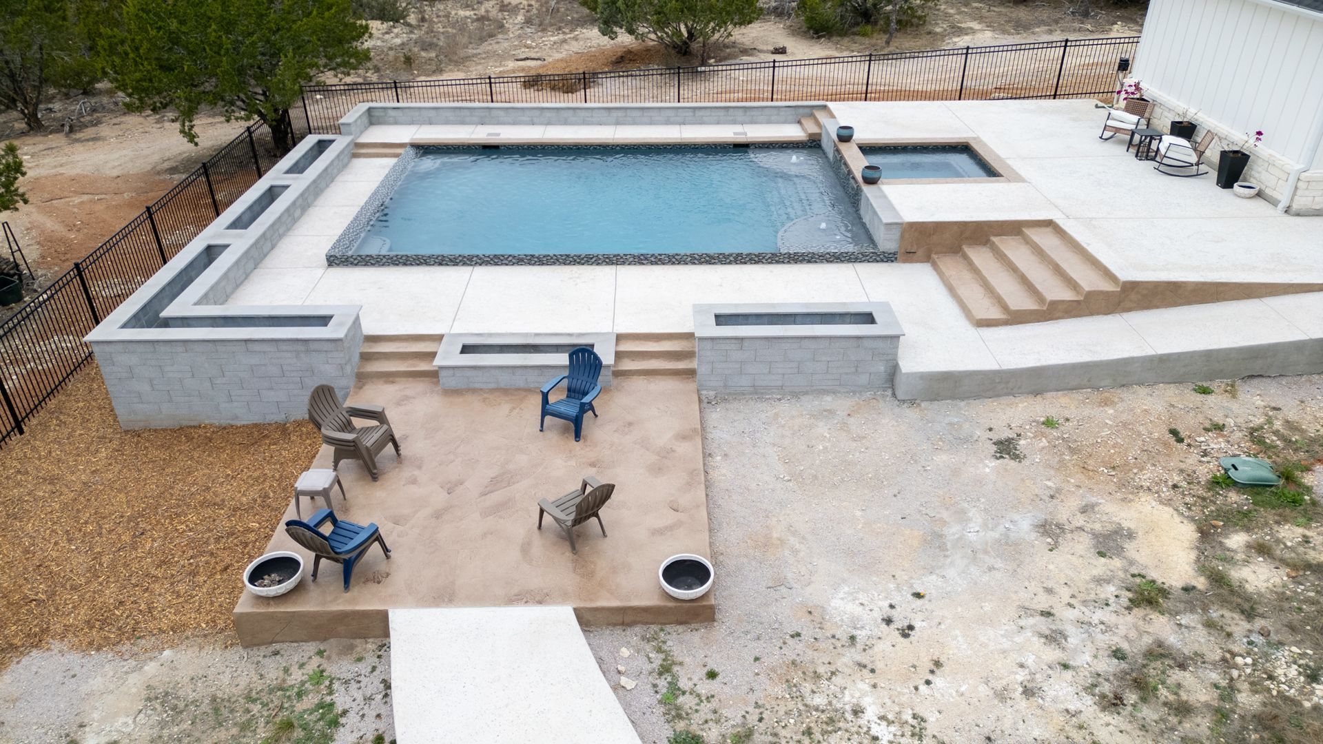 An elevated view of a rectangular backyard pool with an adjacent spa, concrete decking, and a lower seating area with chairs.