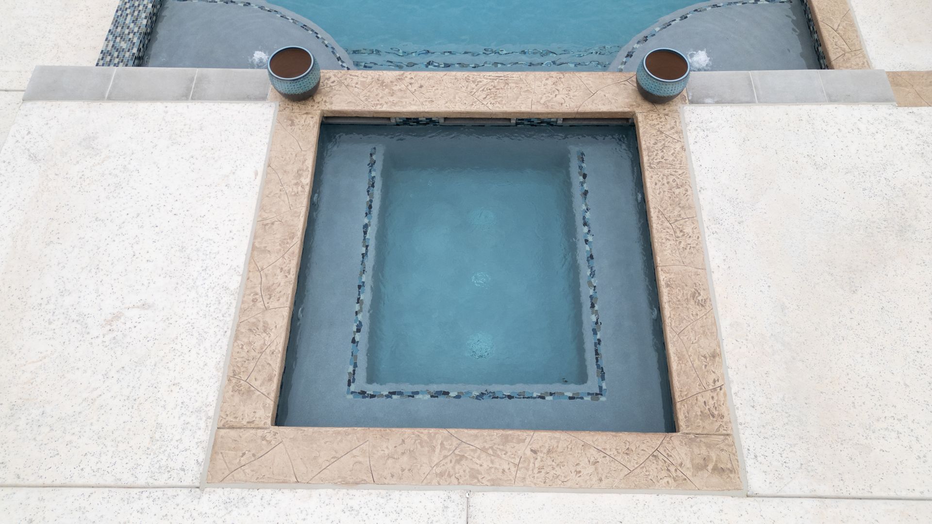 A rectangular spa or pool insert with blue water, set within light-colored stone tiling and two dark decorative vases.