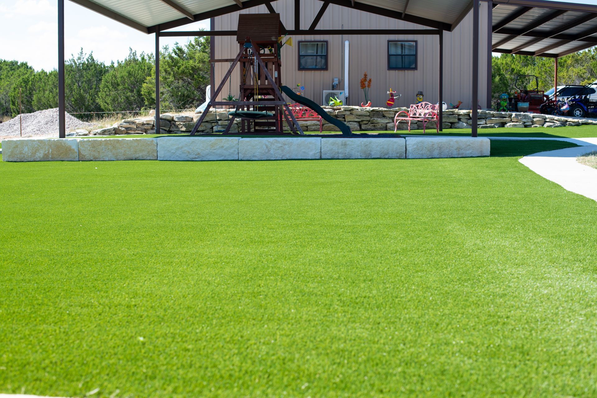 A backyard with artificial green turf, stone retaining walls, and a metal-roofed patio area featuring a wooden playset.