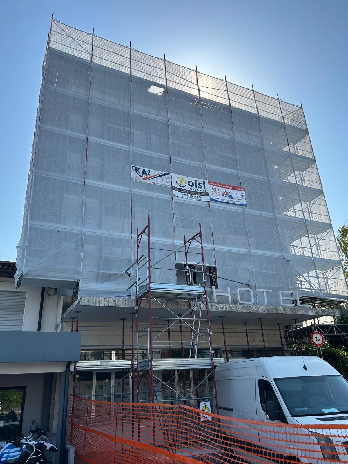 Ponteggi e reti di protezione coprono un edificio in ristrutturazione, con un furgone bianco parcheggiato davanti.