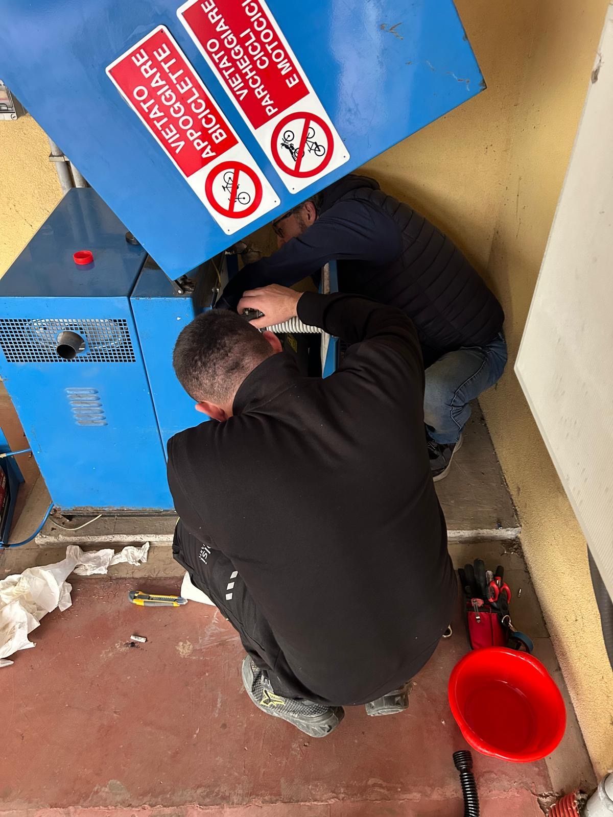 Due operai sono accovacciati accanto a una macchina blu, ispezionandone il pannello inferiore in uno stretto spazio interno.
