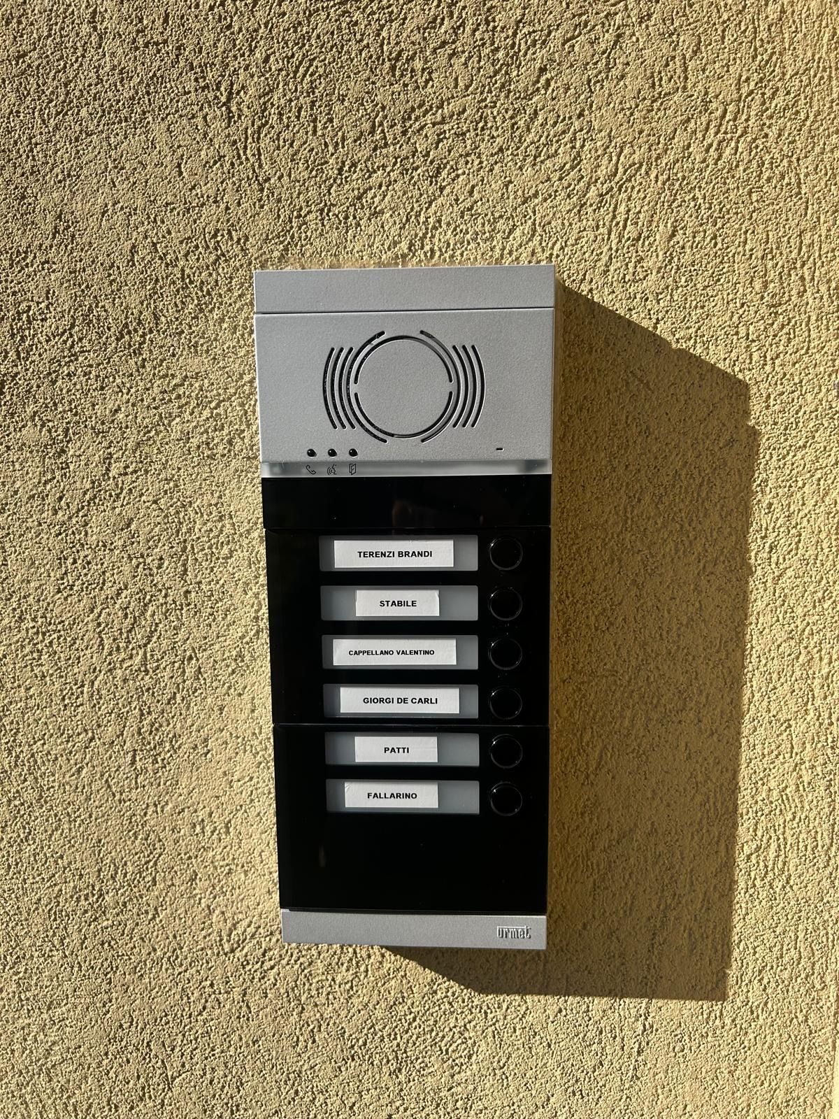 Citofono da appartamento a parete, grigio e nero, con pulsanti etichettati su una parete beige con finitura ruvida.