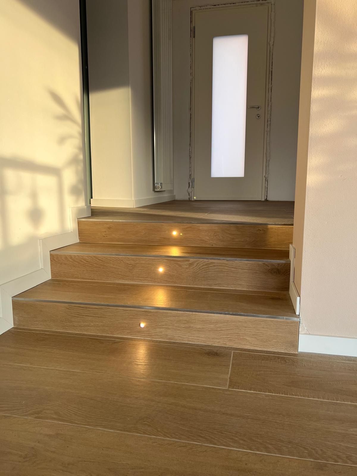 Corridoio interno illuminato dal sole con scalini in legno che conducono a una porta bianca con pannello in vetro satinato.