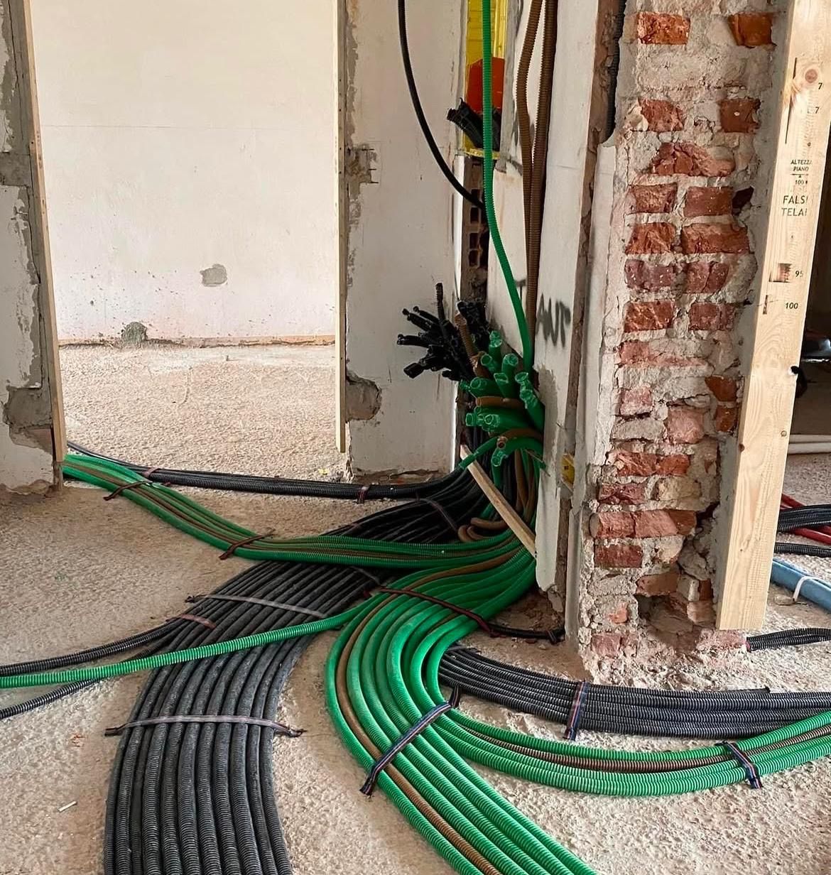 Cavi verdi e neri posati sul pavimento di un cantiere edile, accanto a muri di mattoni a vista e porte.