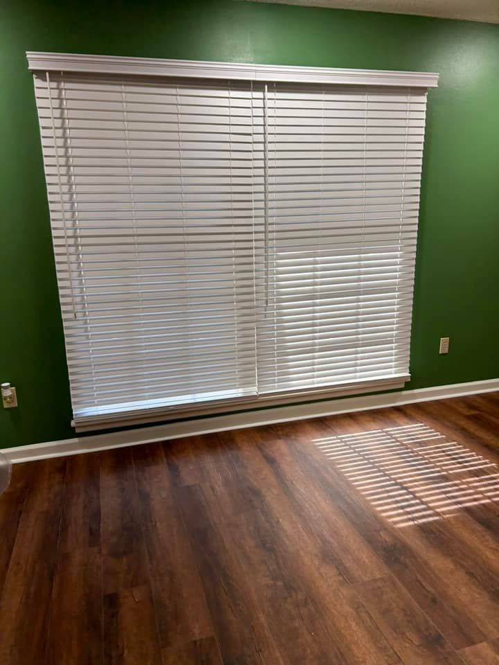 A living room with green walls , hardwood floors , and a large window with blinds.