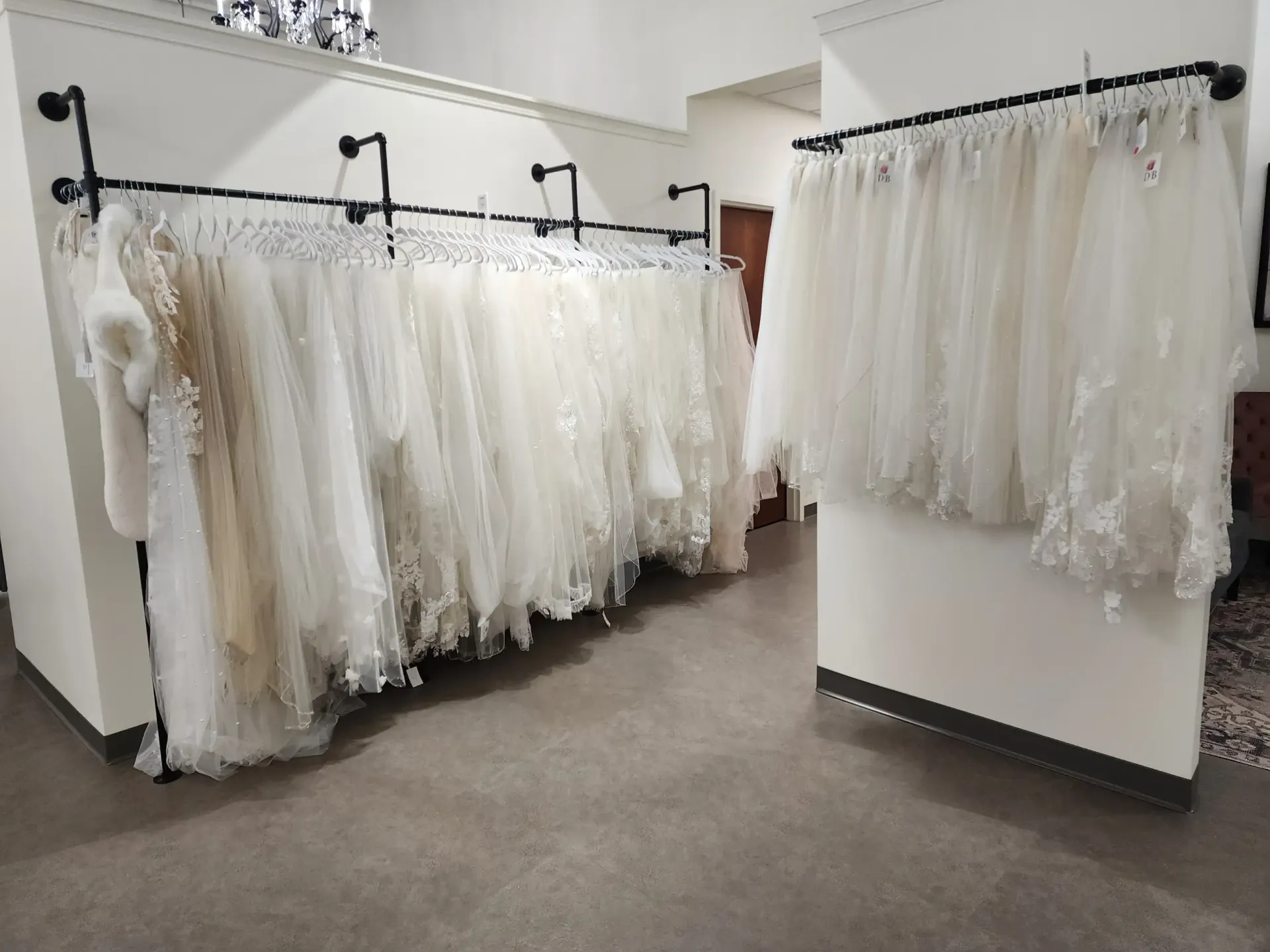 Wedding dresses hang on racks in a bridal shop, in front of a white wall and a brown door.