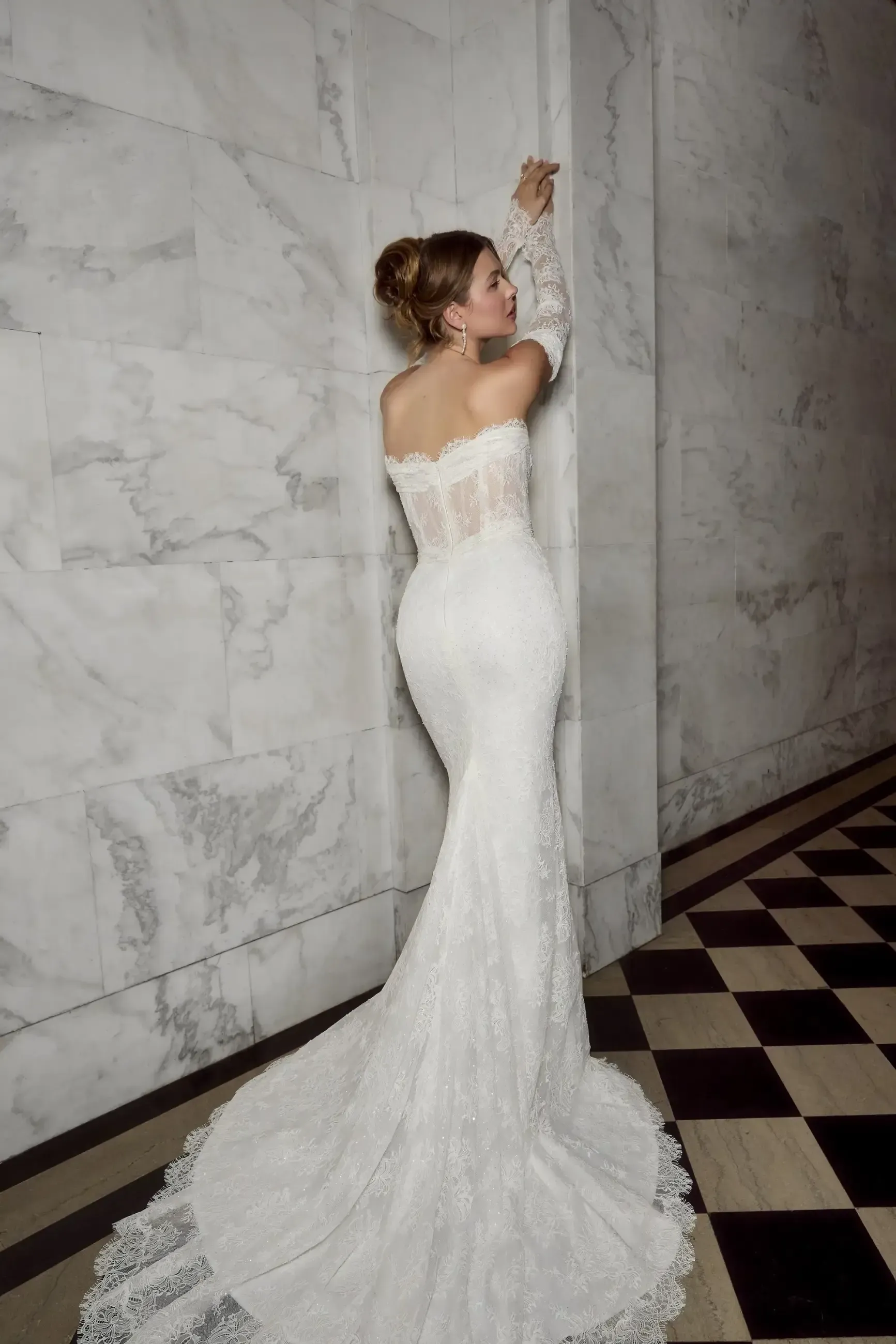 Woman in white, off-shoulder lace wedding dress, poses against a marble wall; long train, updo hairstyle.