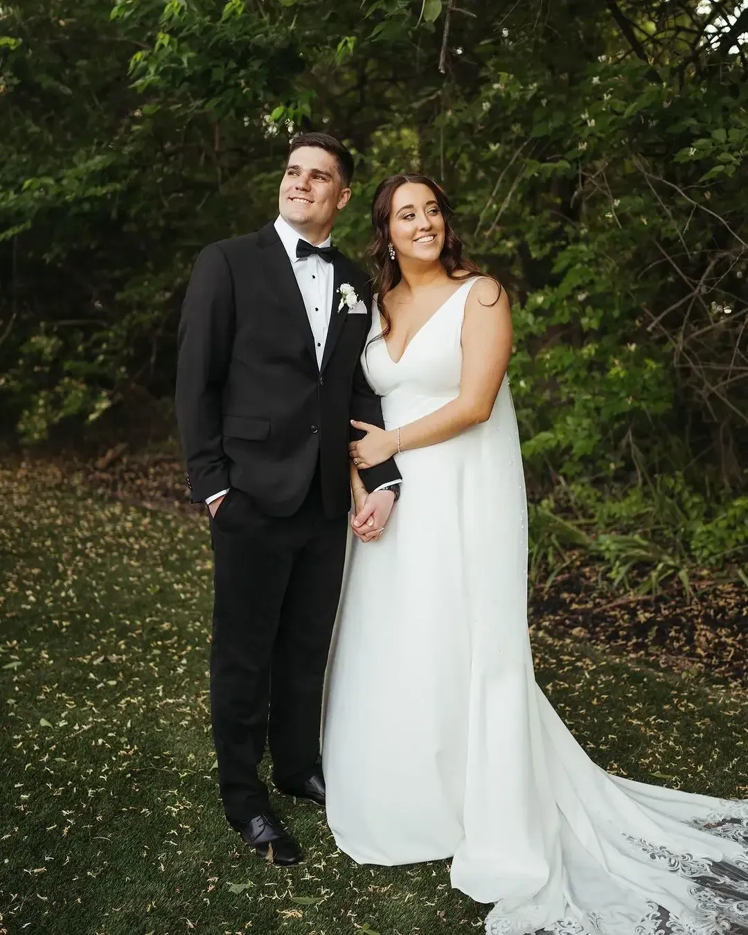 Bride and groom pose outdoors. Man in black suit and bow tie, woman in white gown, holding hands and smiling.