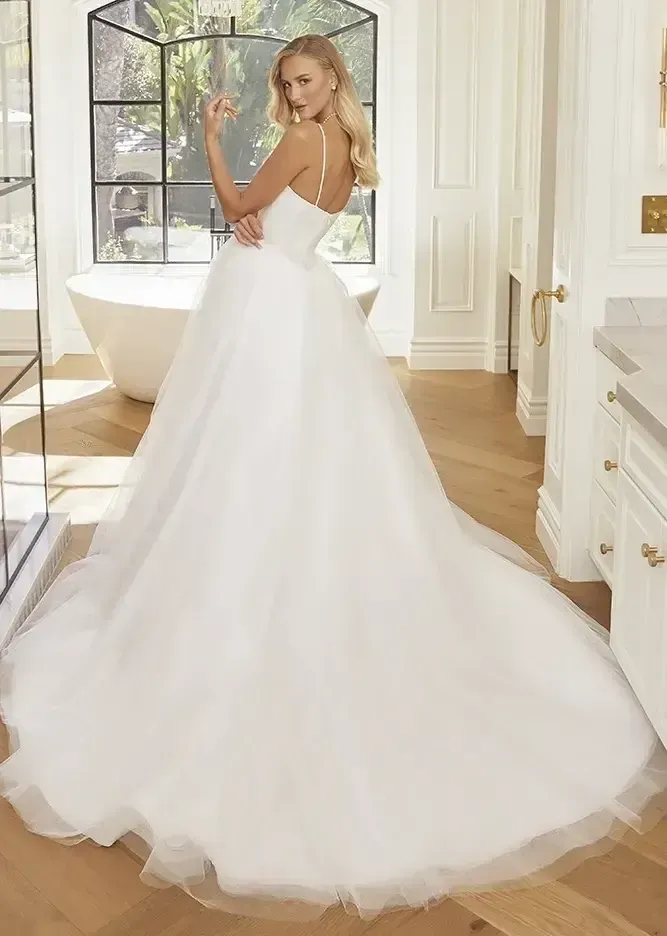 Woman in a white wedding gown with a long train, standing in a bathroom with a large window.