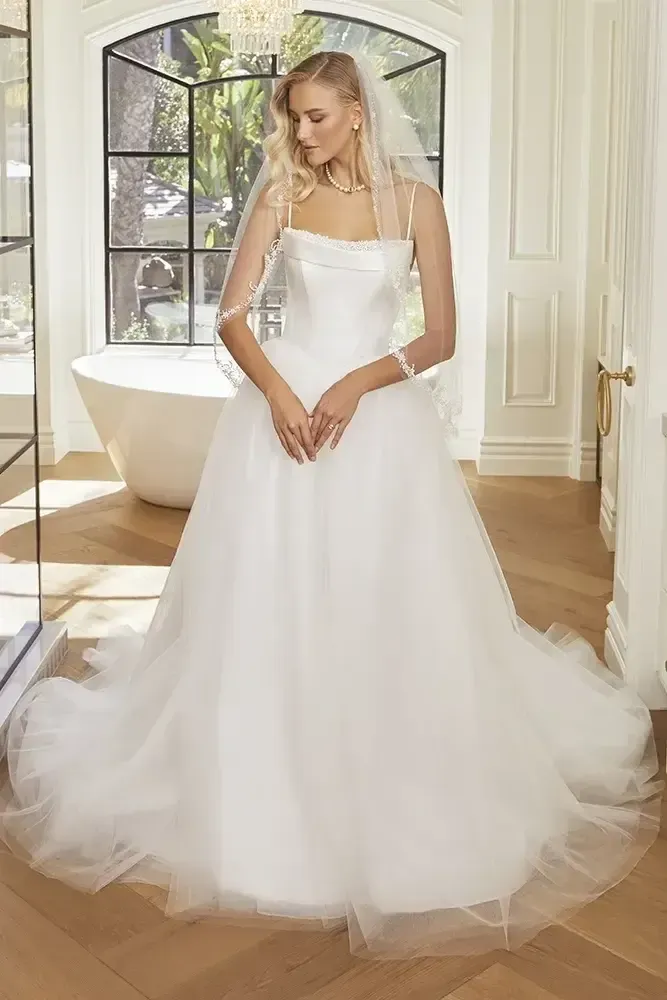 Woman in a white strapless wedding dress, veil, standing in a bright room with a large window and bathtub.