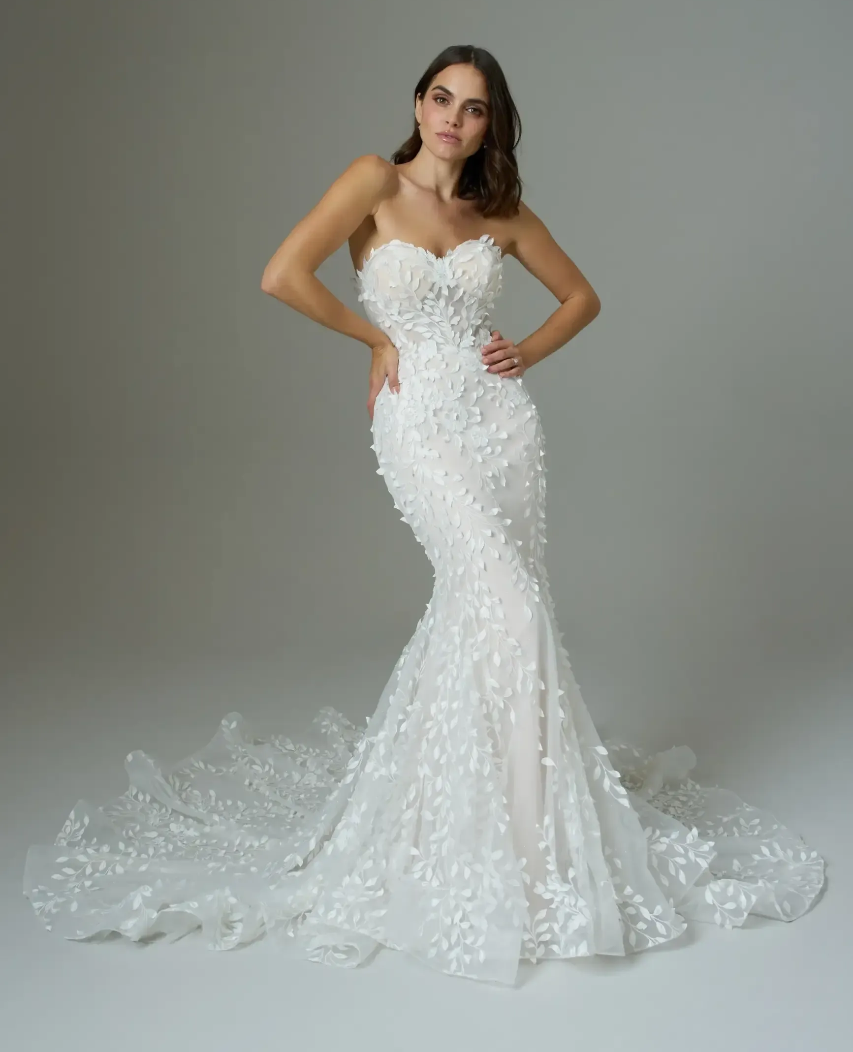 Woman in a strapless white mermaid gown with floral embellishments and long train, posing.