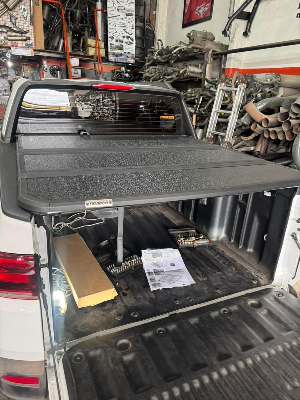 Pickup truck bed with a black tonneau cover, parked inside a cluttered auto shop with the tailgate open