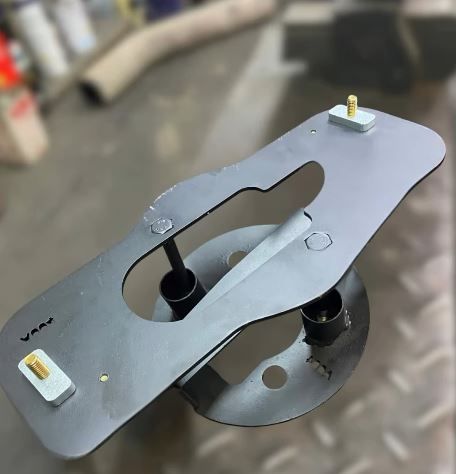 Placa de montaje de metal gris con orificios recortados sobre una superficie de taller.