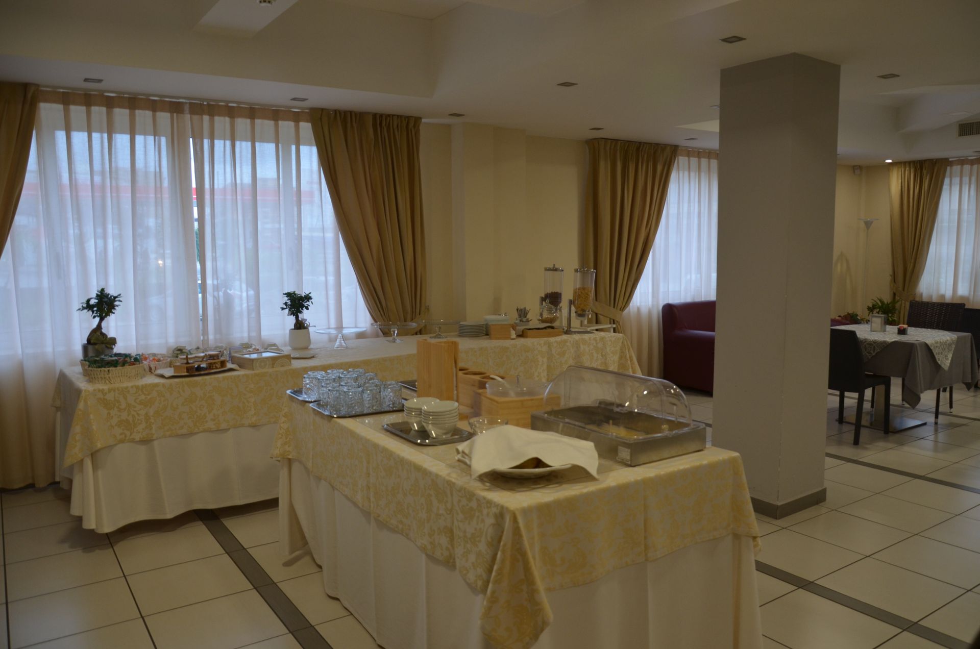 Tavoli da buffet con cibo nella sala da pranzo di un hotel; arredamento beige e bianco, tende.