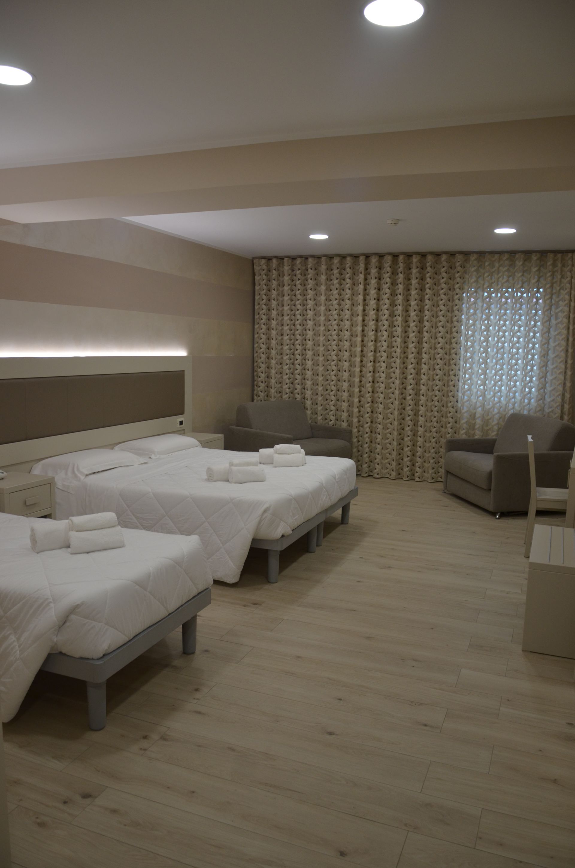 Camera d'albergo con tre letti, due poltrone e una finestra con tende a fantasia. Tonalità beige e legno chiaro.