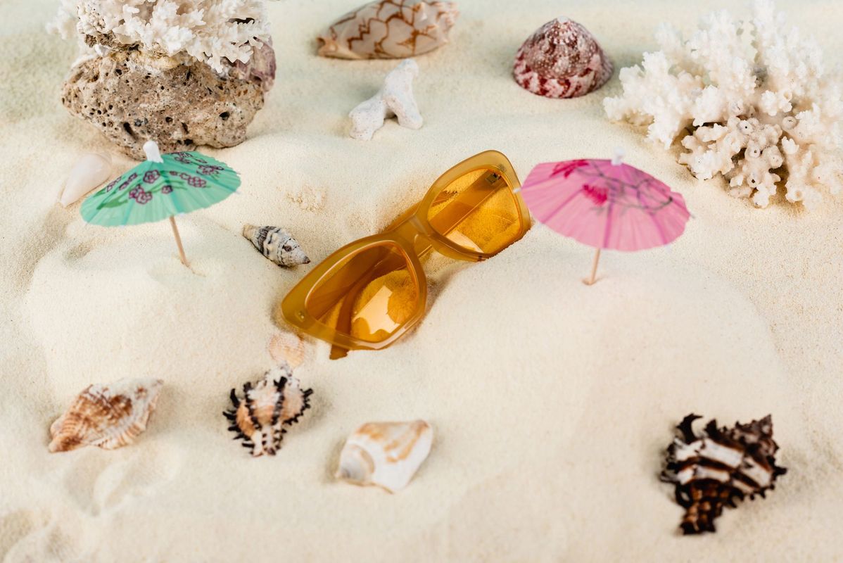 Occhiali da sole su una spiaggia sabbiosa con conchiglie e ombrellini da cocktail.