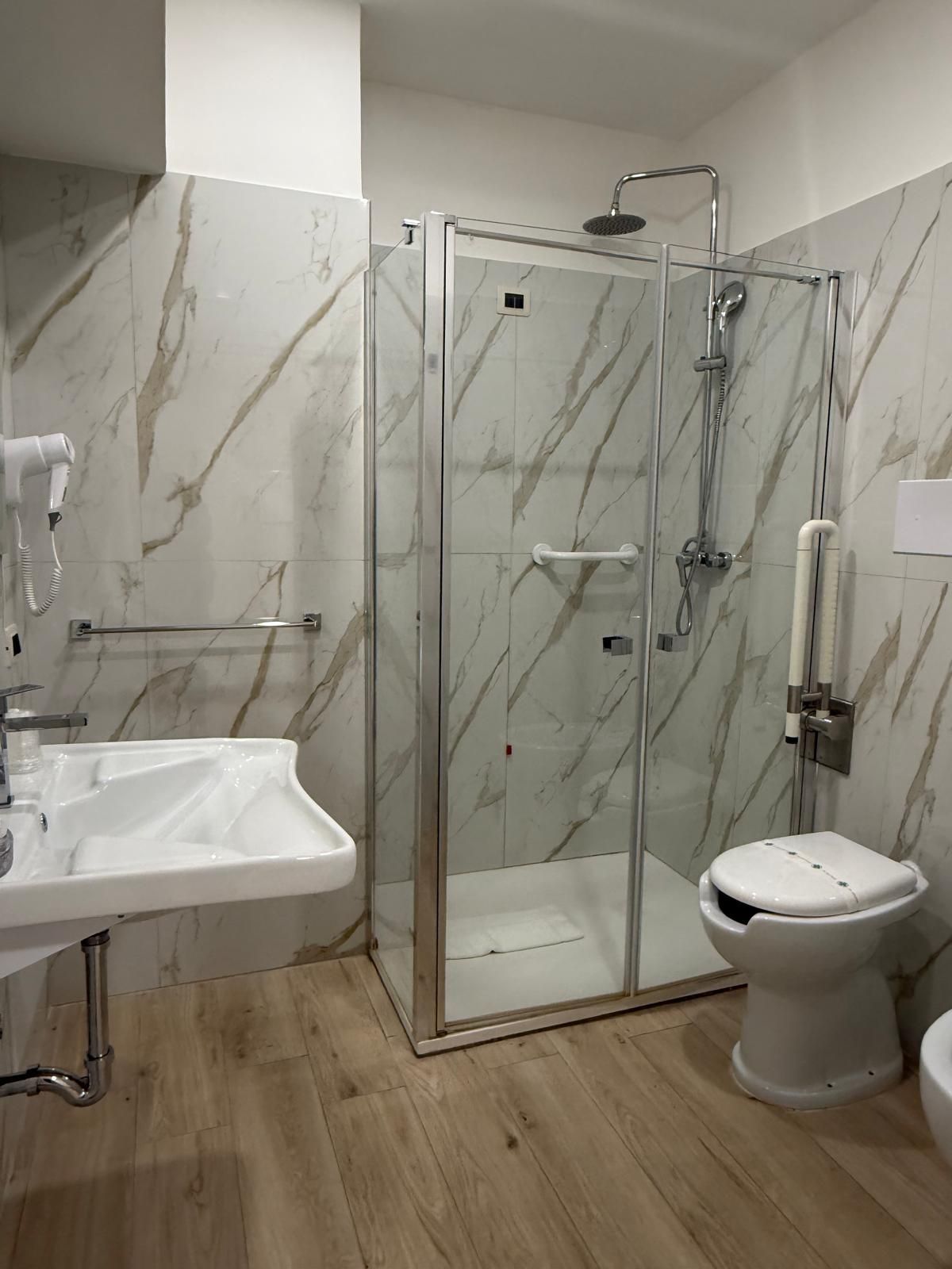 Bagno con doccia in vetro, WC e lavandino, pareti con motivi in marmo e pavimento effetto legno.