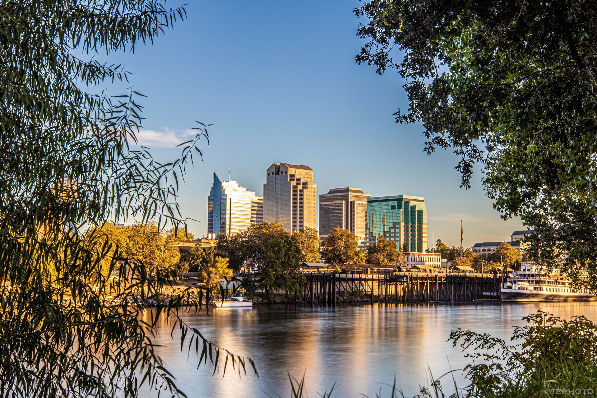 City skyline reflected on calm water, framed by trees at sunset.
