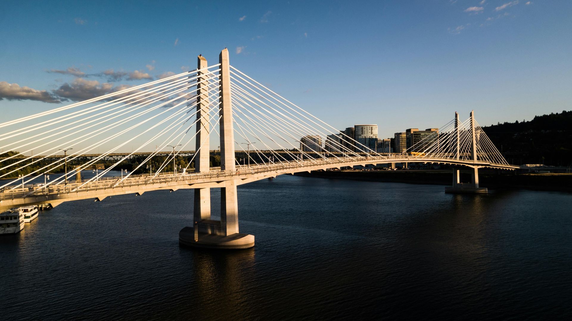 Cable-stayed bridge over a wide river at sunset, with bright cables and calm water