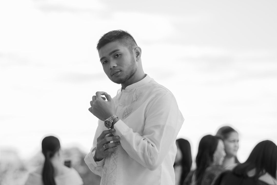 Black-and-white portrait of a man in a white shirt, posing with a hand near his chin outdoors, blurred crowd behind him