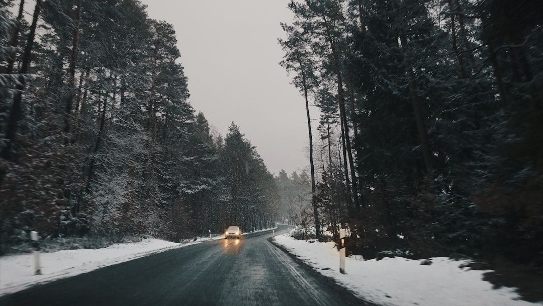 Snowy forest road at dusk with a car driving toward the camera and headlights on.