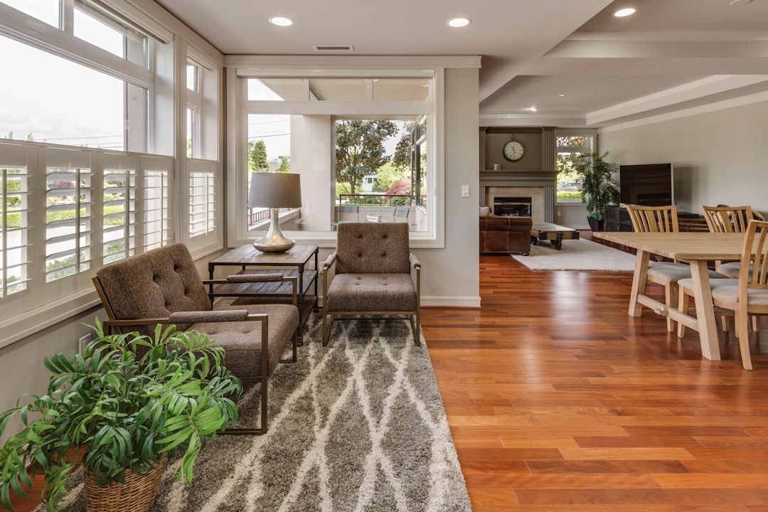 Bright open living room with hardwood floors, tufted chairs, patterned rug, potted plants, and dining area visible.