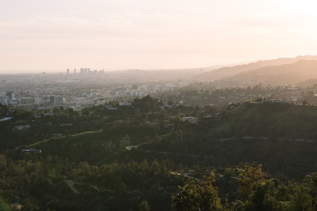 Hazy city skyline beyond green hills at sunrise, with soft pink sky and distant mountains