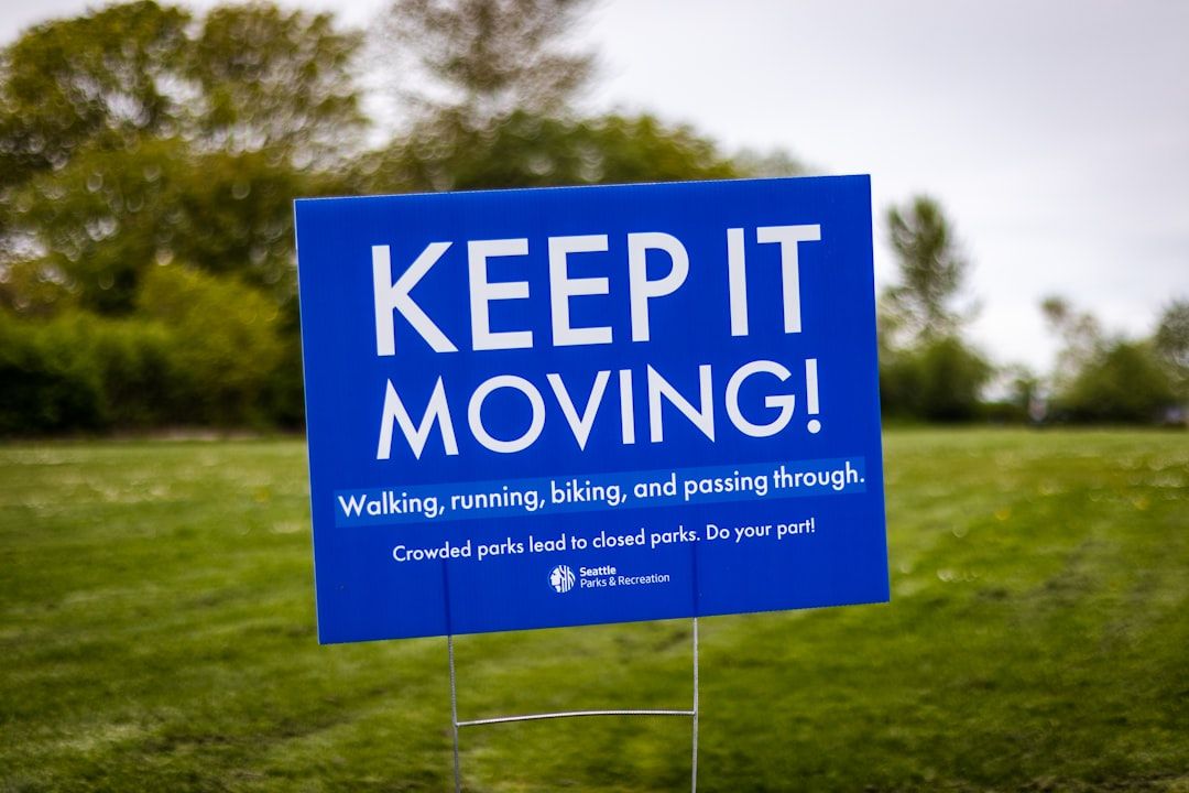 Blue sign in a grassy park reads “KEEP IT MOVING!” with trees in the background.