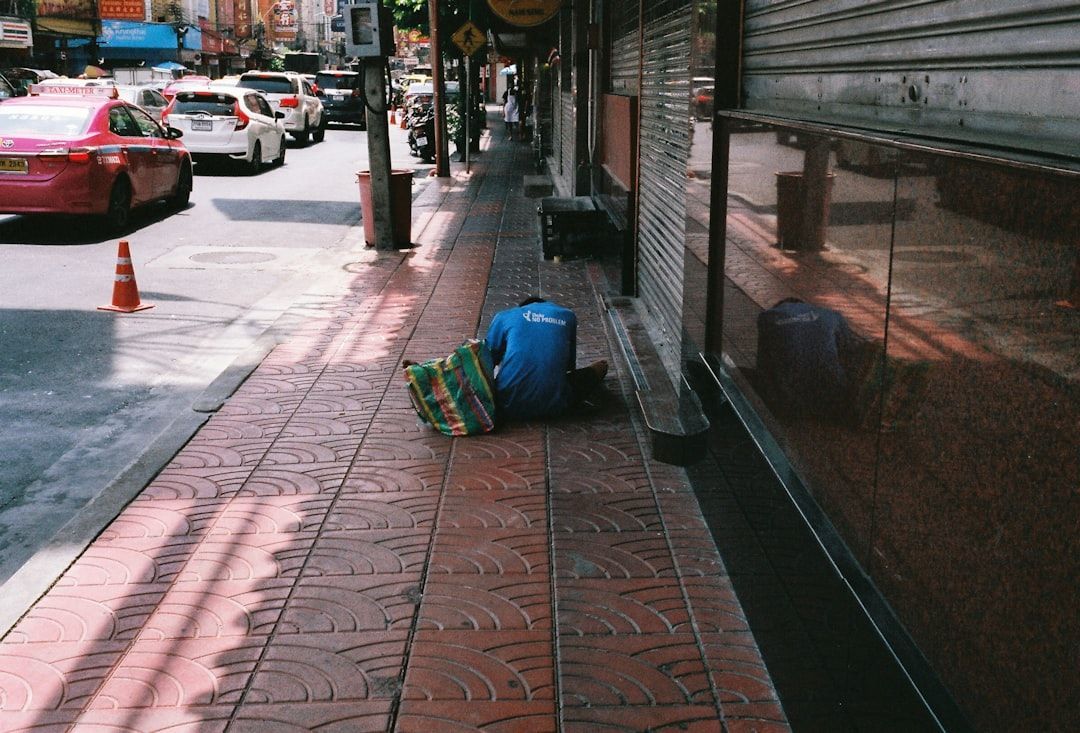 Person crouched on a sidewalk beside a storefront, near a blue bag on a rainy city street