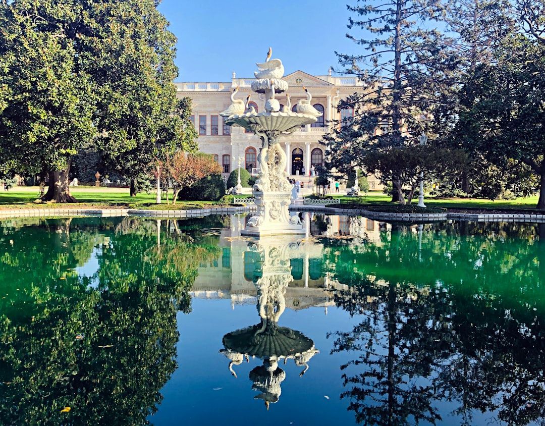 Fountain reflecting a grand white building in a tree-lined garden with a green pond