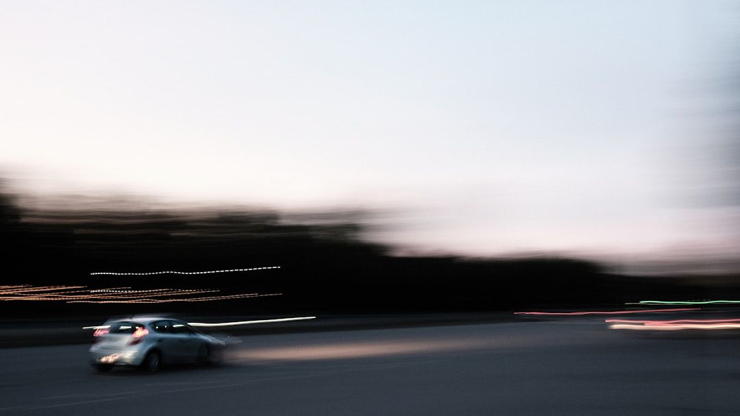 Blurred white car speeding across an open road at dusk with dark trees in the background
