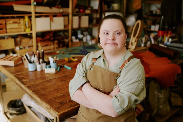 Person standing with arms crossed in a woodshop, wearing an apron, with tools and shelves in the background