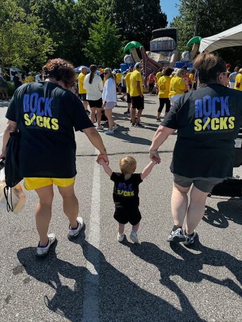 Crowd walking outdoors; two adults and a toddler in matching black shirts reading “ROCK STARS” hold hands.
