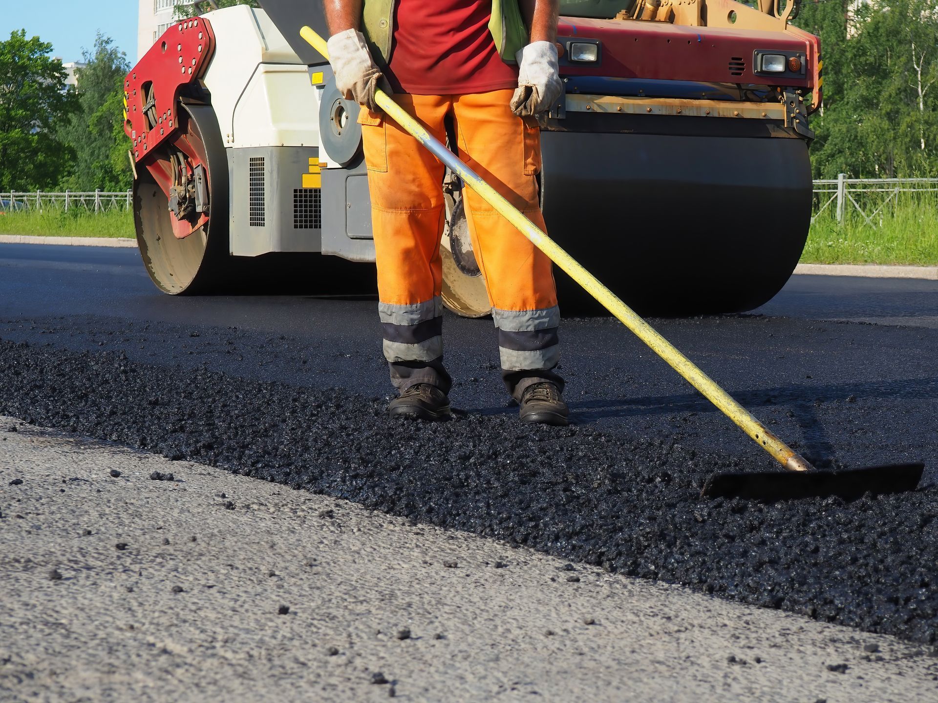 A worker rakes asphalt while a roller compacts pavement near trees and a fence. A worker rakes asphalt while a roller compacts pavement near trees and a fence.