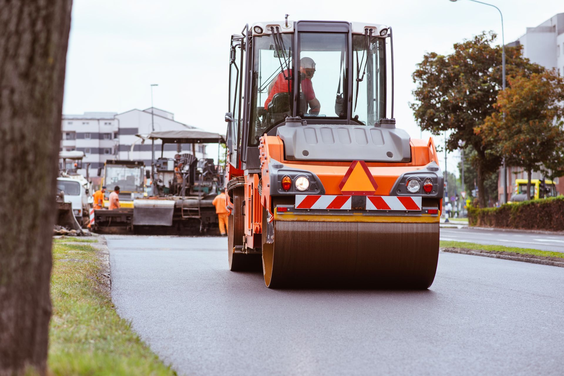 Road roller compacting freshly laid asphalt on a street. Worker in cab; other road work in background.