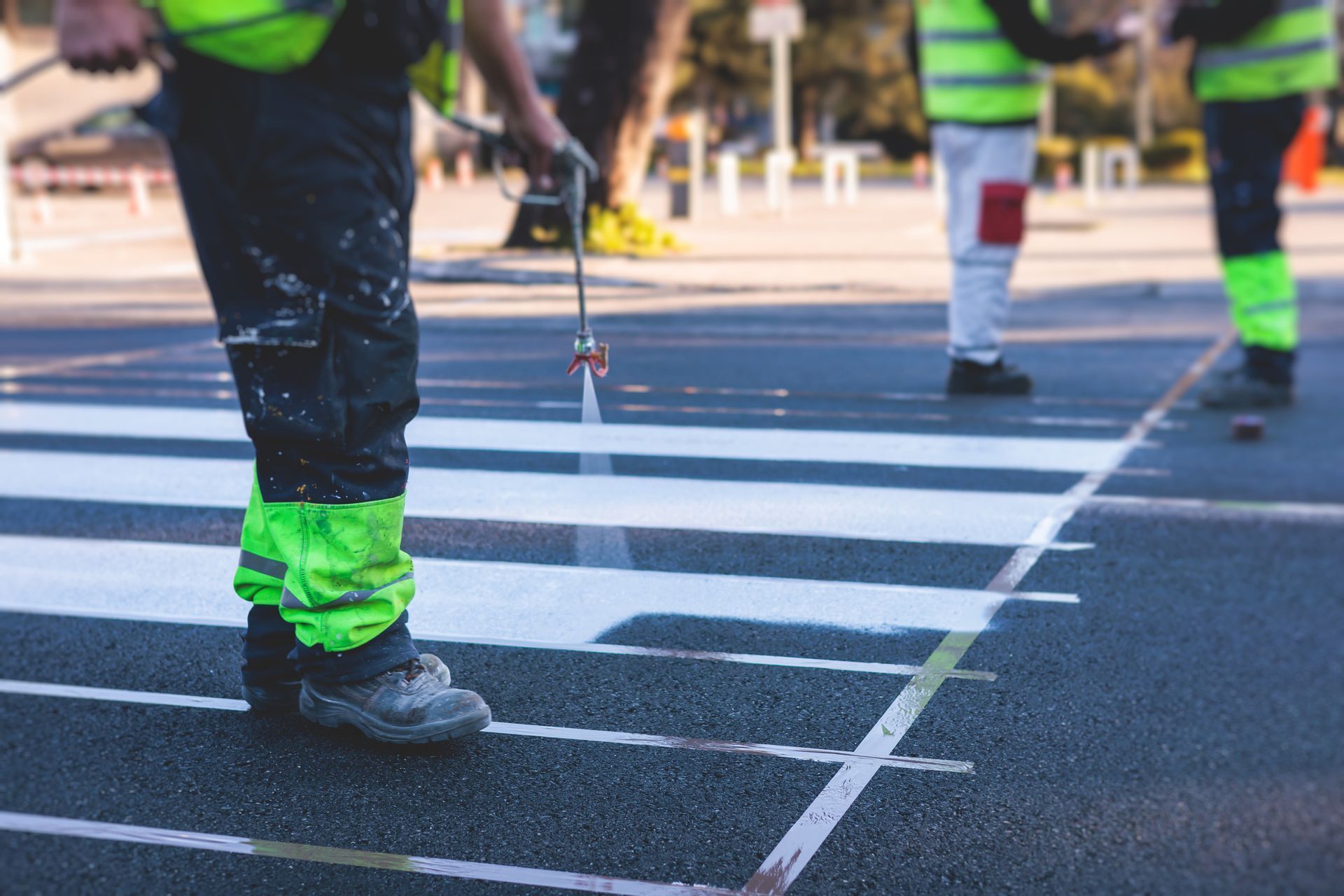 Workers repainting a crosswalk with white paint on a city street, wearing safety vests.