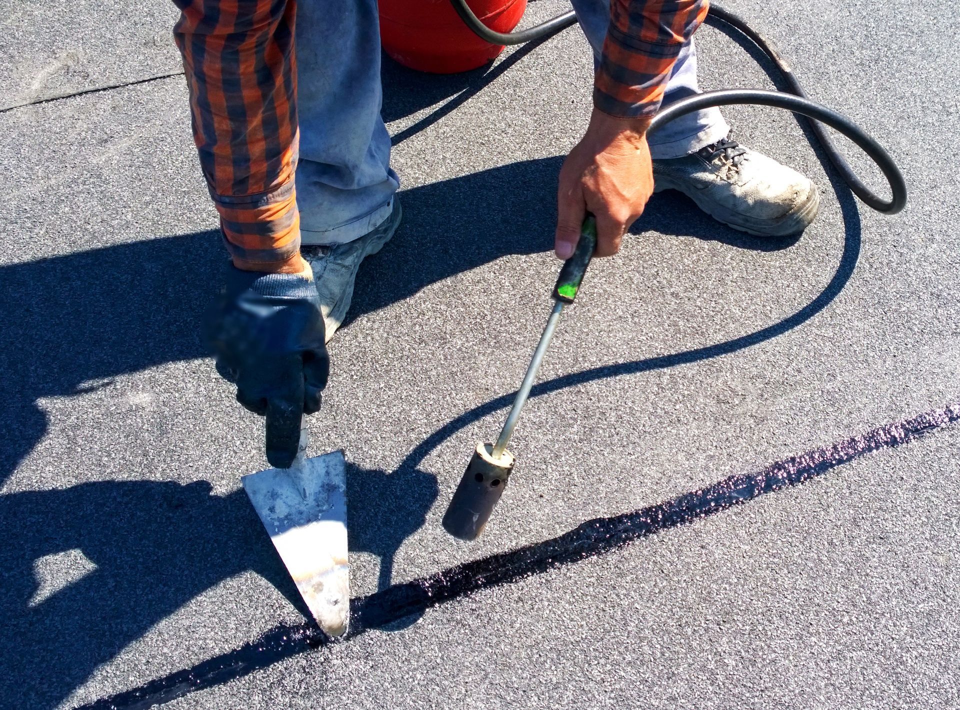 Roofer using a torch and trowel to apply sealant on a dark, granular surface, possibly a roof.