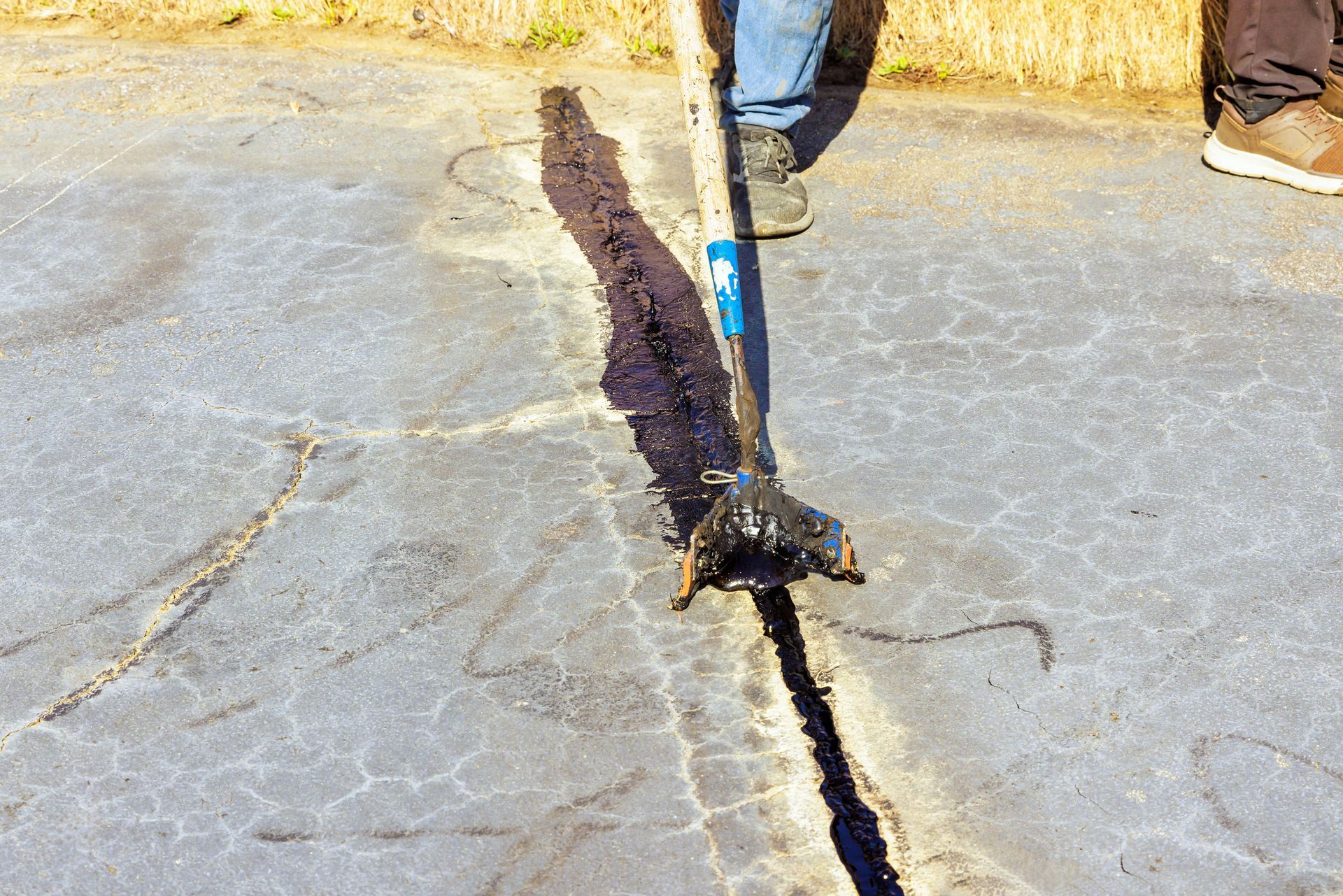 Person using a tool to apply black sealant to cracks in an asphalt surface.