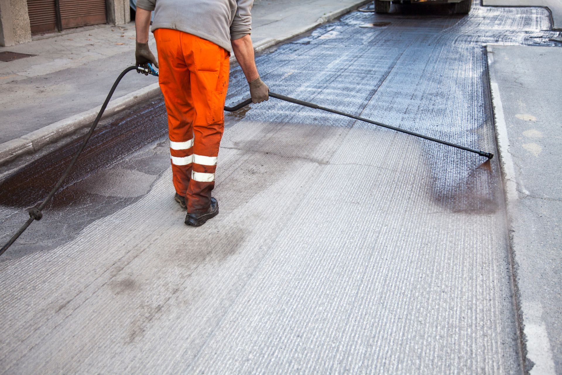 Road worker in orange pants spraying asphalt with a liquid sealant on a street.